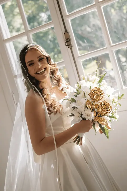 Bride by a window holding a bouquet of white lilies and gold dipped roses, real roses dipped in 24K gold
