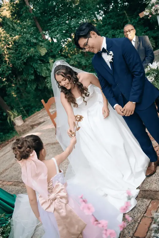 Flower girl adjusting the bride’s dress beside a groom holding a gold dipped rose, gold rose dome gift ideas for Monaco
