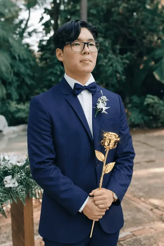 Groom in a navy suit holding a single gold dipped rose at the altar, gift inspiration for the Imperial Majestic Noir gold dipped rose (one real rose preserved in 24K gold, black crocodile leather)