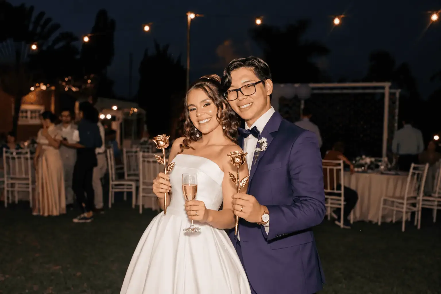 Bride and groom holding gold dipped roses and champagne at an evening wedding reception, a gold dipped rose celebration